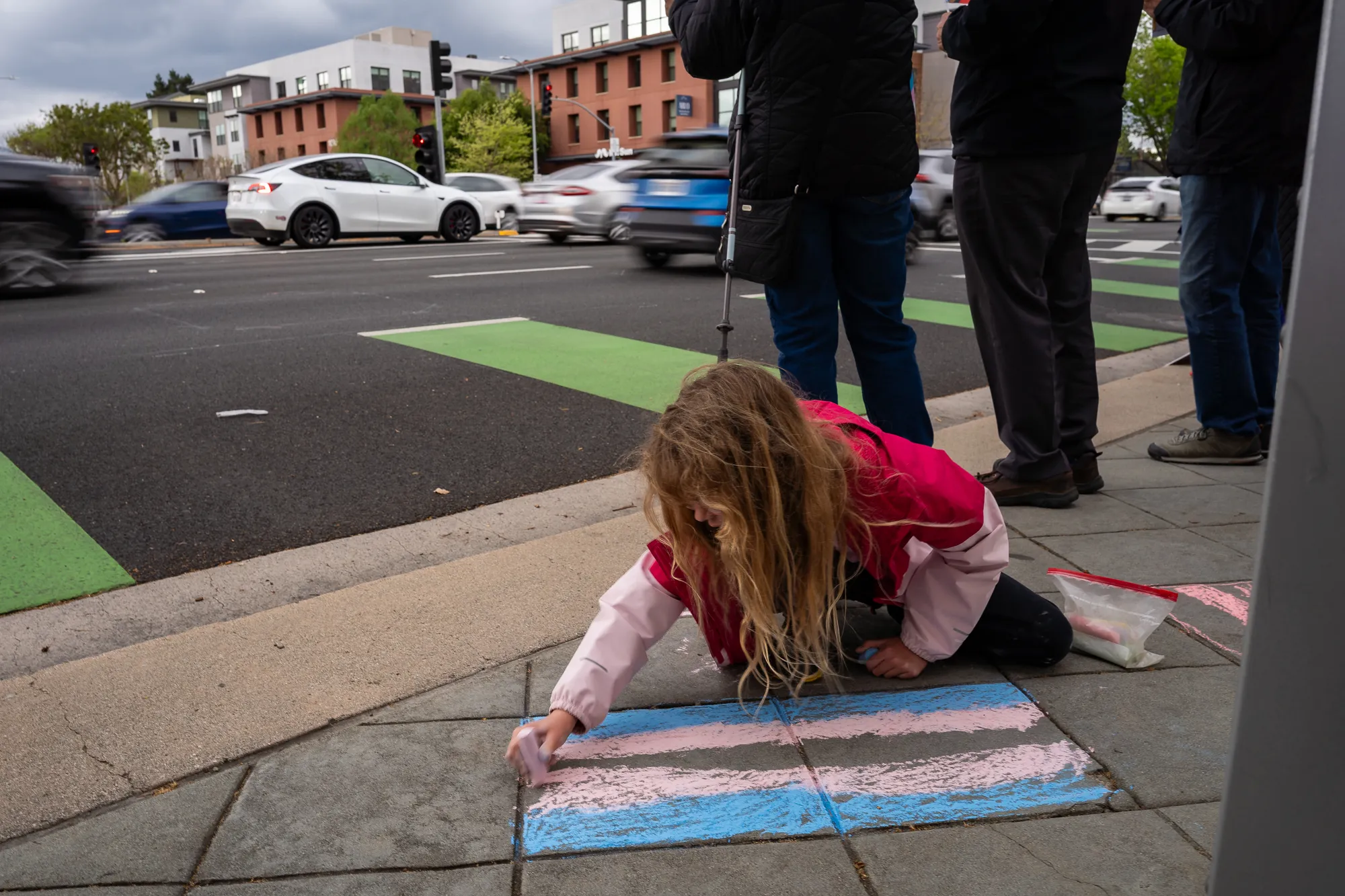 A child draws a trans pride flag in chalk on the sidewalk. Photo by Seeger Gray.